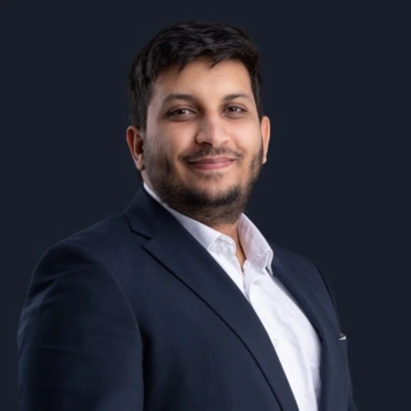 A man with short dark hair and facial hair wearing a navy blazer and white shirt, smiling against a dark background.
