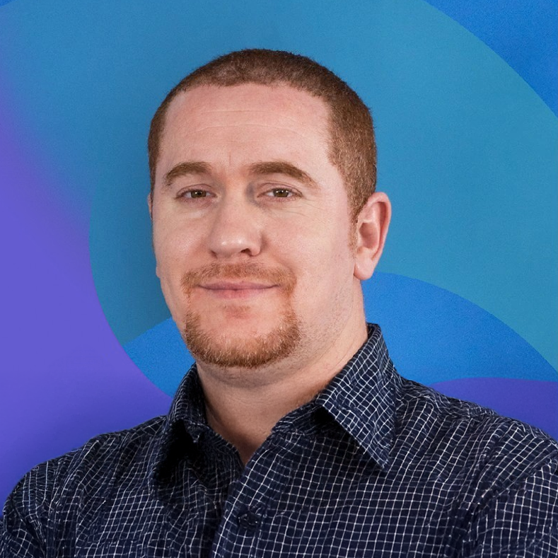 A man with short red hair and a goatee wearing a dark checked shirt, standing in front of a blue and purple abstract background.