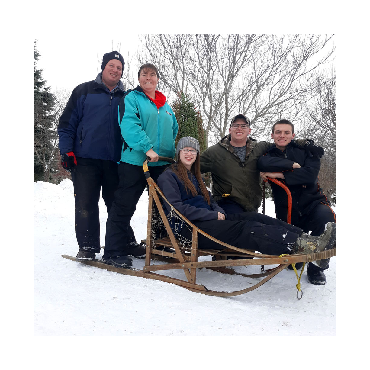 Five people on a snow-covered hill with a sled. Two men are standing behind, one woman and two young men are sitting on the sled. They are all dressed in winter clothing, and there are snow-covered trees in the background.