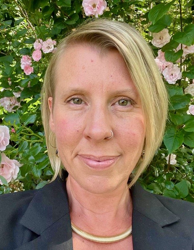 A woman with short blonde hair, light skin, wearing a black blazer, gold necklace, and earrings, standing in front of green leafy bushes with pink and white flowers, smiling.
