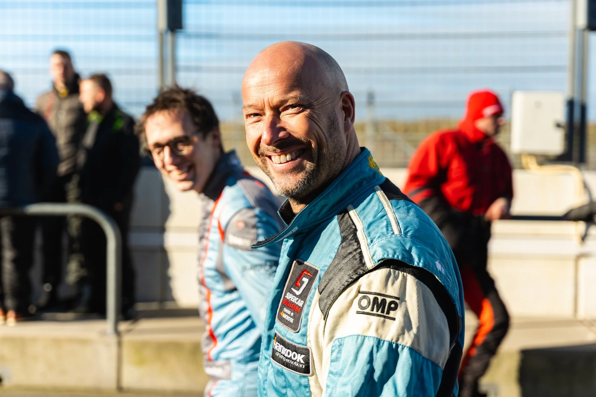 Smiling race car drivers standing on a race track, with one man in the foreground wearing a racing suit with various sponsor patches, and a woman and other people in the background.