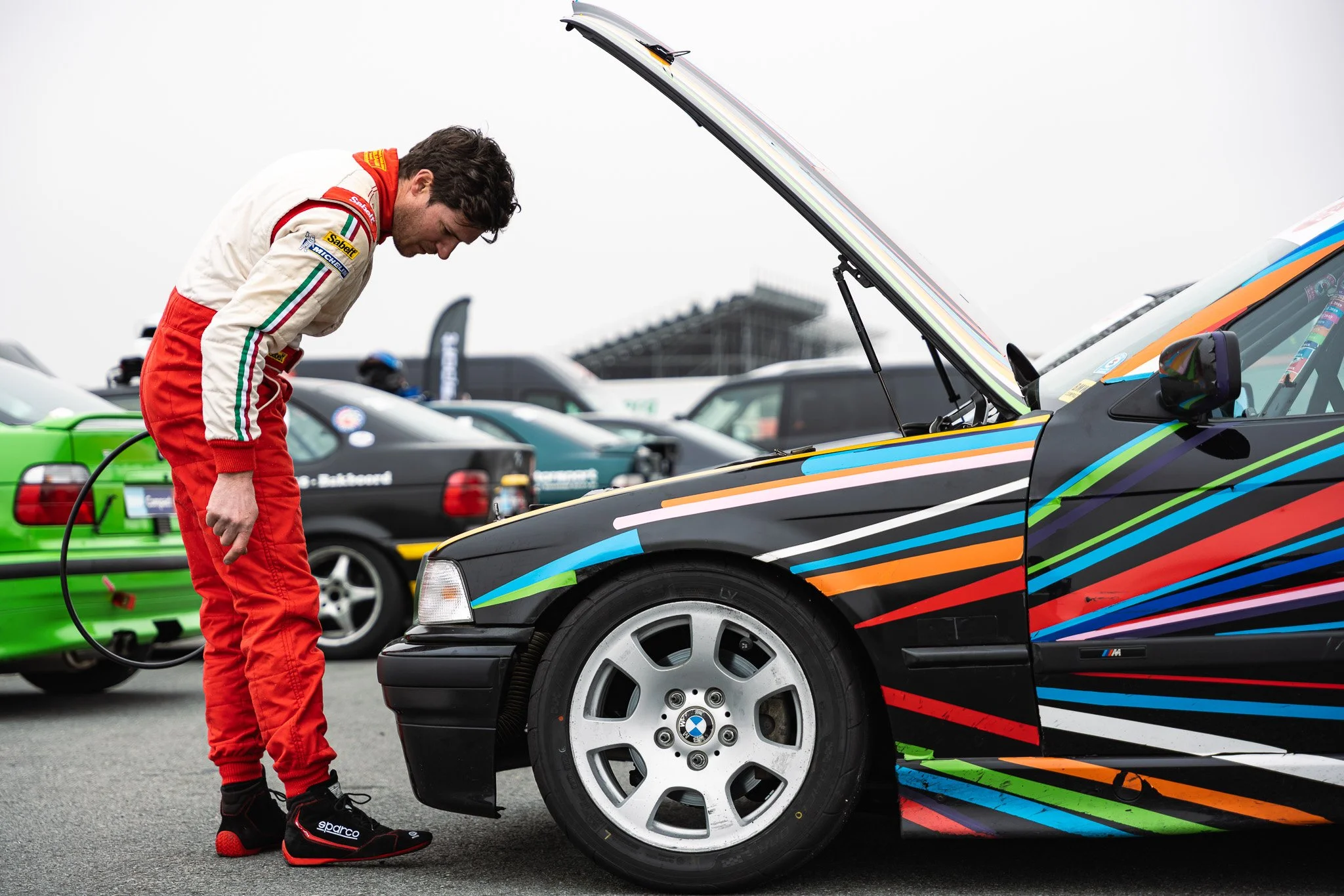A race car driver in a red and white racing suit stands next to a black race car with colorful stripes and an open hood, examining or working on the vehicle at a race track.