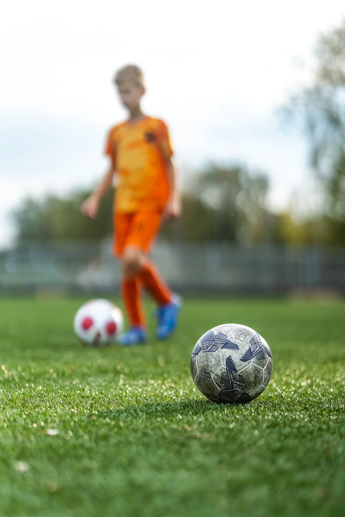 Een kind in een oranje sportoutfit staat op een grasveld met twee voetbalen, één voor en één achter hem, met een achtergrond van bomen en een heldere lucht.