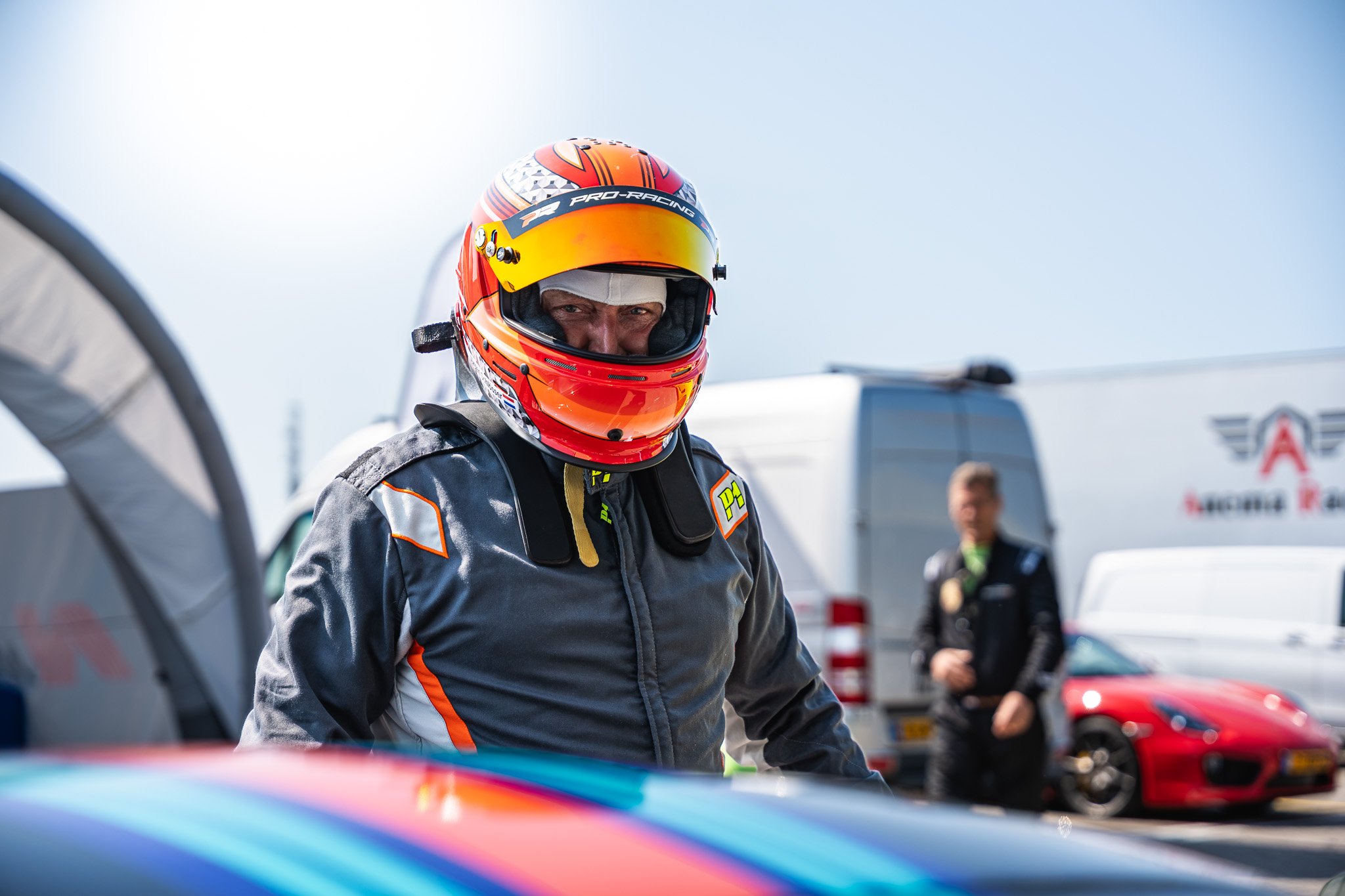 Een racecoureur met een rood oranje helm kijkt aandachtig. Hij draagt een zwart en grijs racesuit met oranje accenten en staat tussen racewagens en vrachtwagens op een circuit.