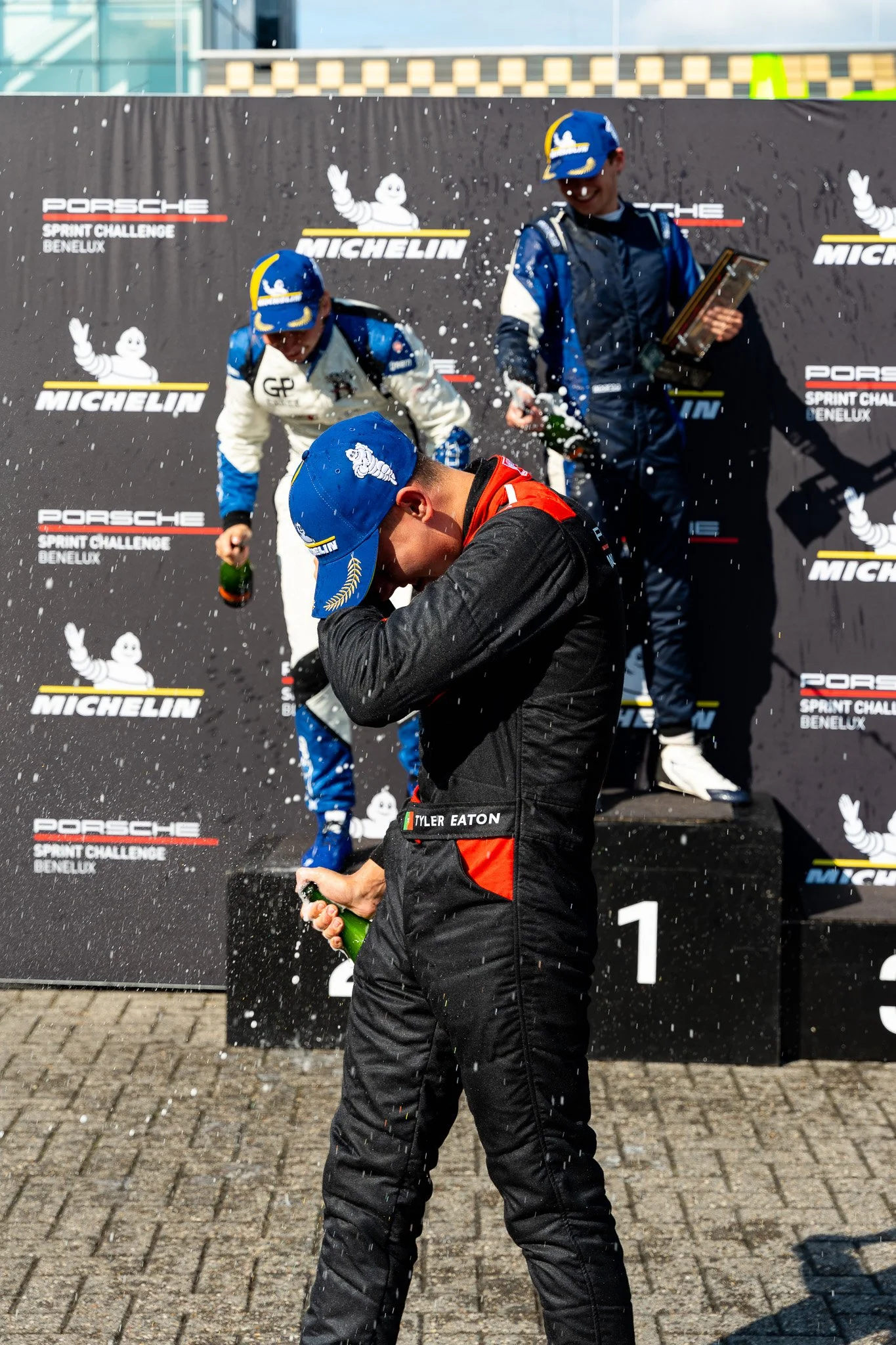 Winnaars op het podium van de Porsche Sprint Challenge Benelux vieren hun overwinning met champagne, met vaten en confetti op de achtergrond.