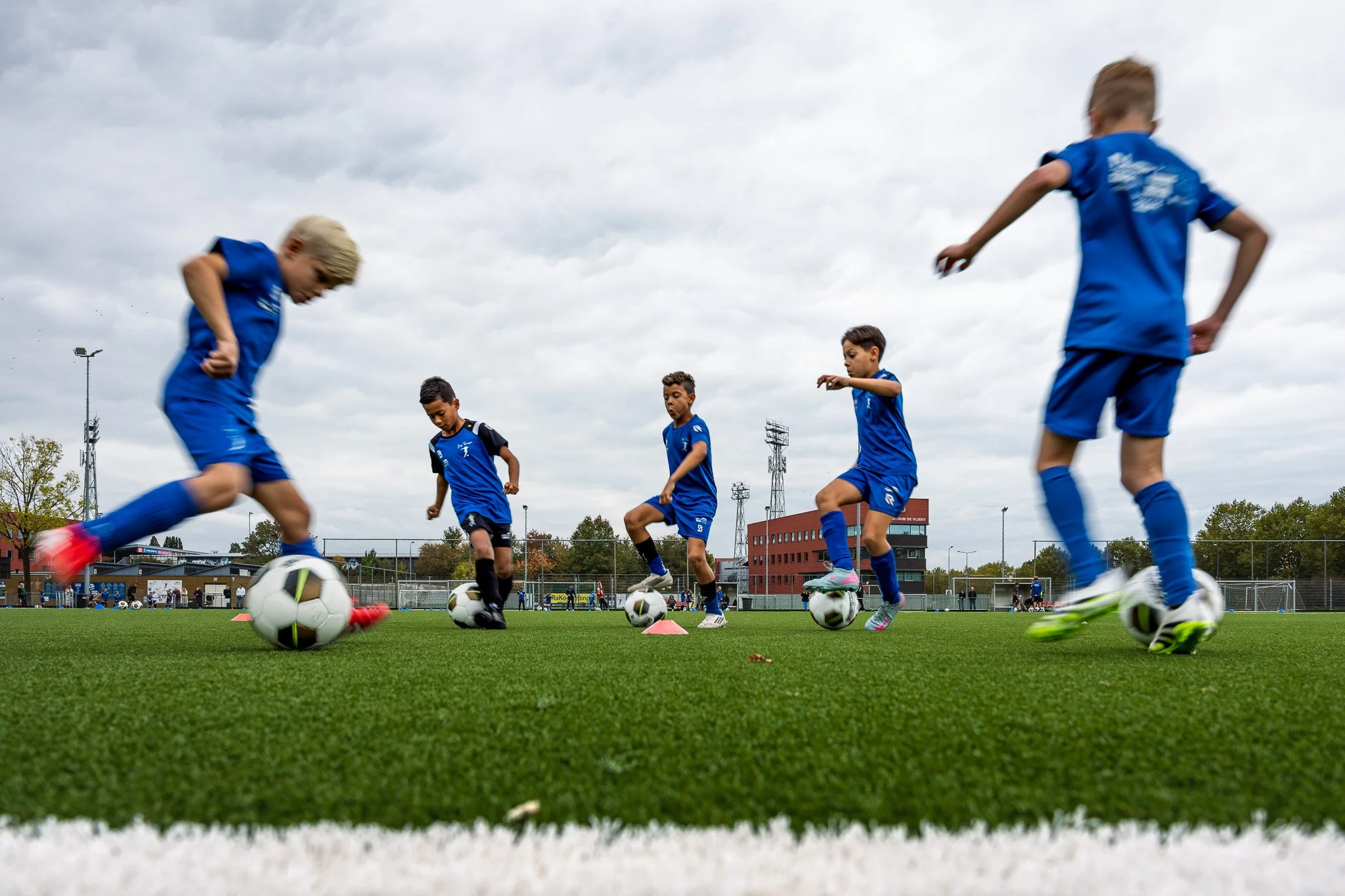 Jongens spelen in een voetbaltraining op een grasveld, allemaal dragen blauwe sportkleding en voetbalschoenen, met voetballen en pionnen op het veld, onder een bewolkte hemel.