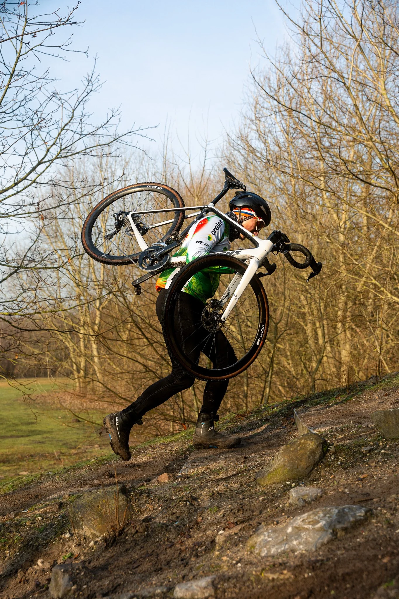 A cyclist carrying a bicycle uphill on a dirt trail in a wooded area during daytime.