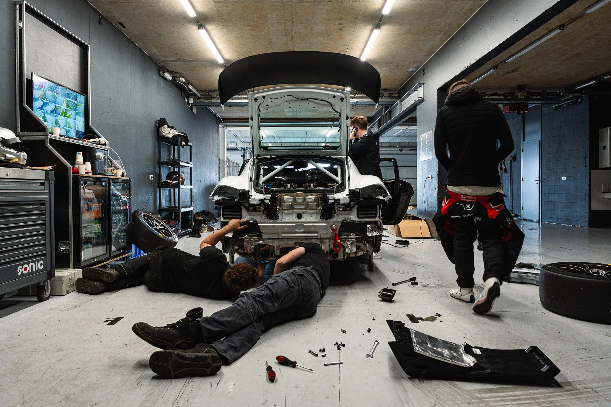 Mechanics working on a race car in a garage, with tools scattered on the floor and the car's hood open.
