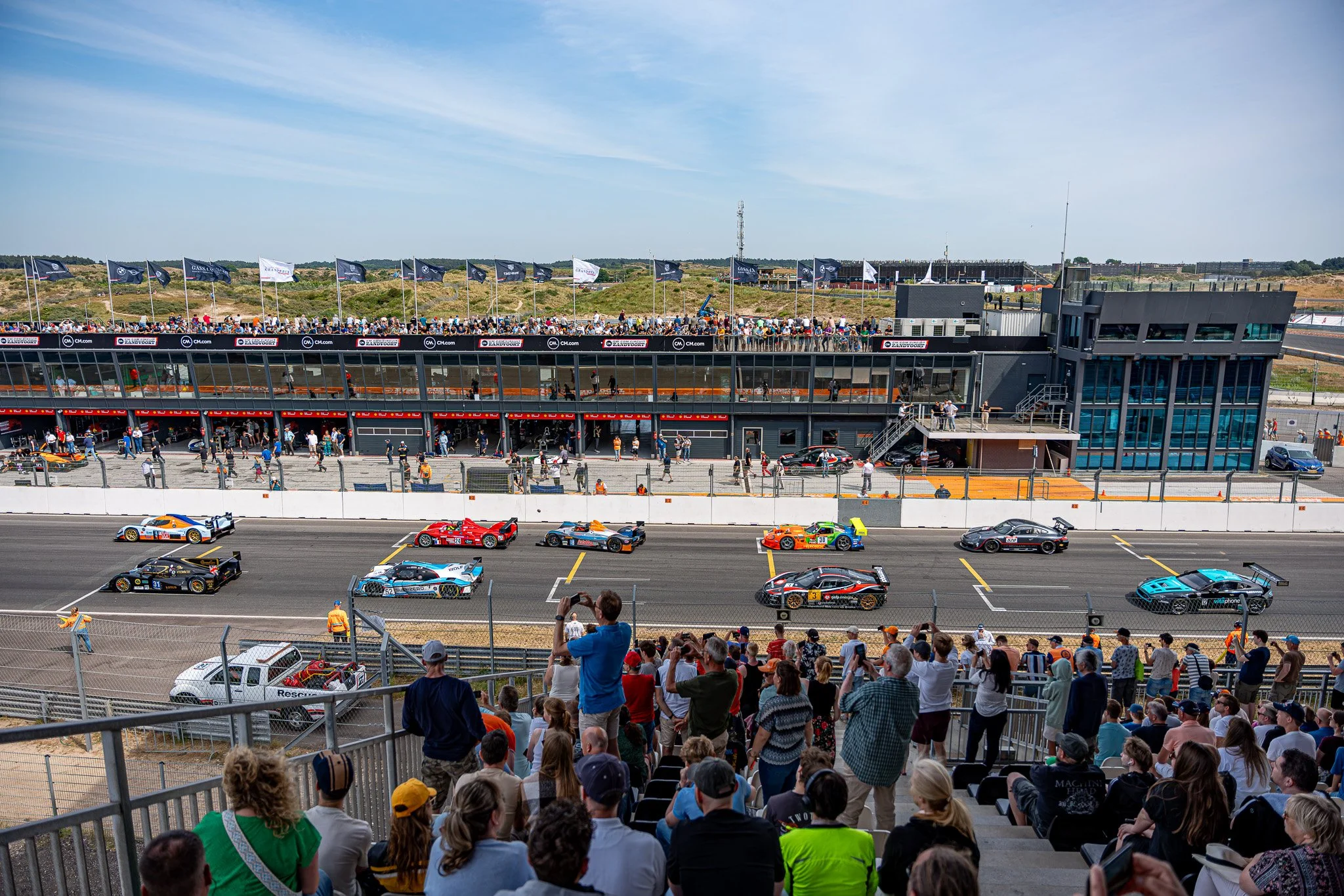 Race fans kijken vanaf een tribune naar een autosportwedstrijd met verschillende raceauto's op het circuit, vlaggen wapperen in de wind op de achtergrond onder een heldere blauwe hemel.