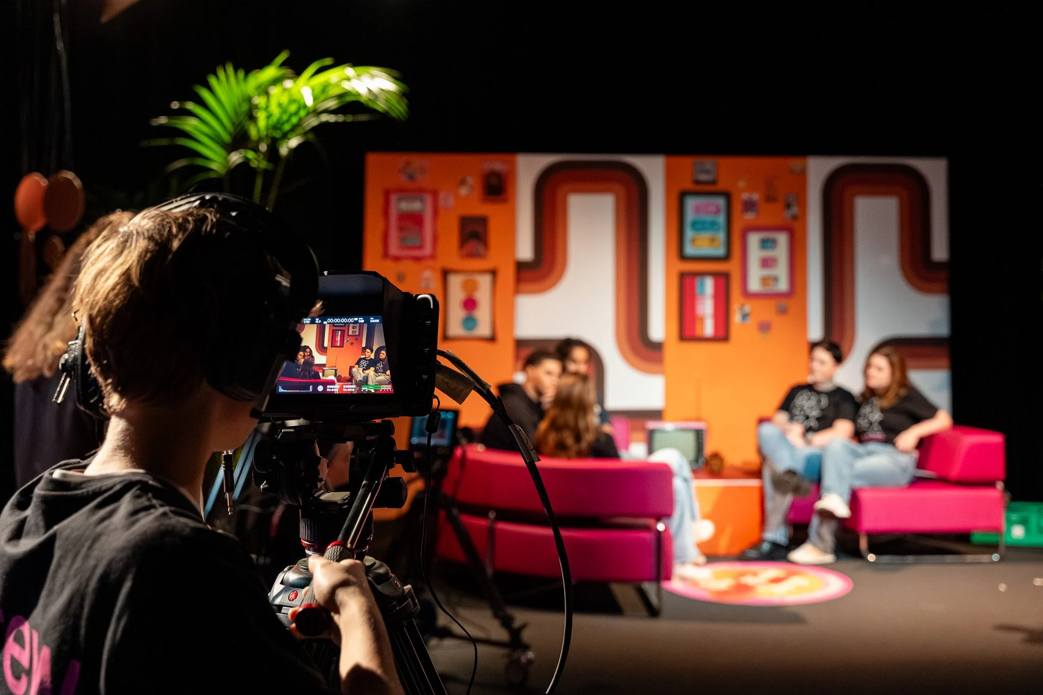 Person filming a group of people on a pink couch in a colorful, retro-style setting.