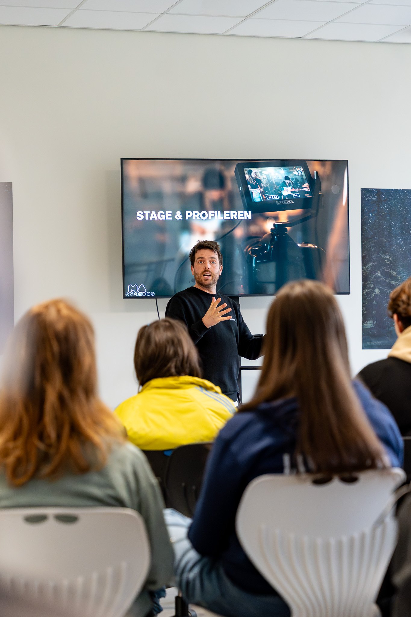 A man giving a presentation to a group of people in a classroom setting. The presentation slide on the screen behind him reads "STAGE & PROFILEN" and shows a camera filming a performer.