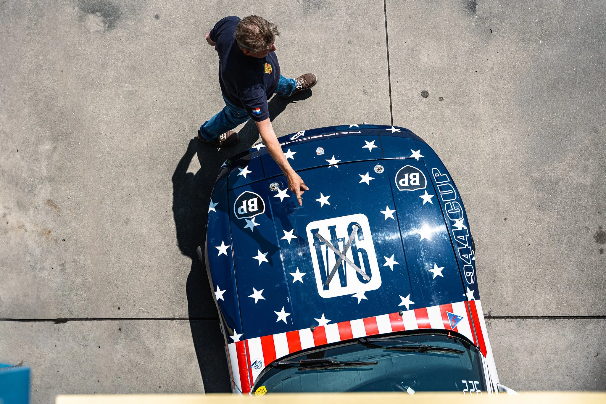 Een van bovenaf genomen foto van een man die op een parkeerplaats voor een raceauto staat, de auto heeft een rode, witte en blauwe Amerikaanse vlagstijl met sterren en strepen. De man draagt een donkerblauw shirt met een NL-vlag en een badge, en is a