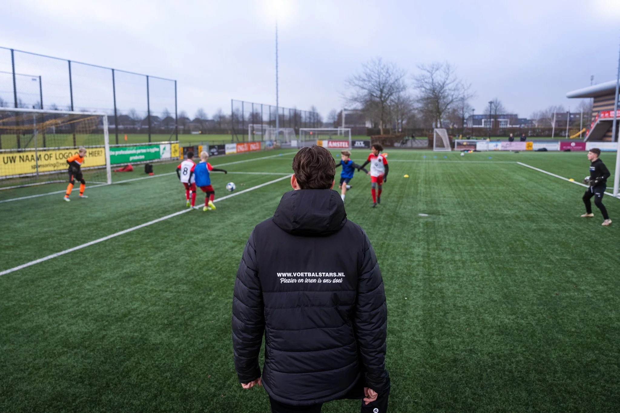 Bekijk van achteren een persoon in een zwarte jas die toekijkt hoe jonge kinderen op een voetbalveld spelen, met doelen en omheining op de achtergrond.