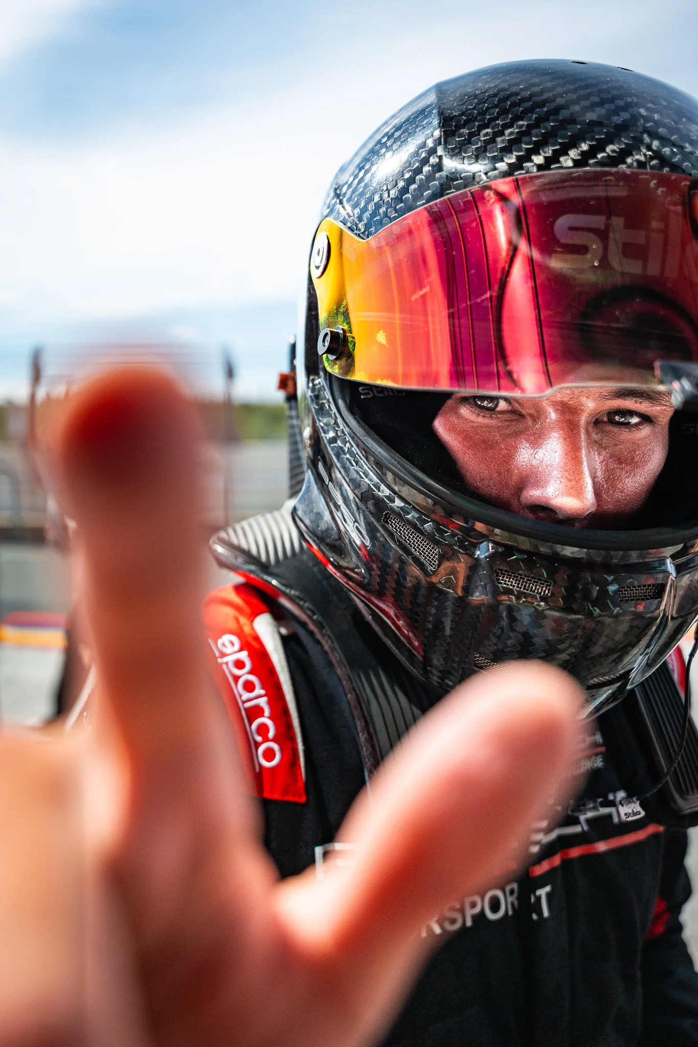 A race car driver wearing a black helmet with a red visor and a racing suit is reaching out towards the camera with an open hand. The background is blurred, focusing on the driver's face and helmet.