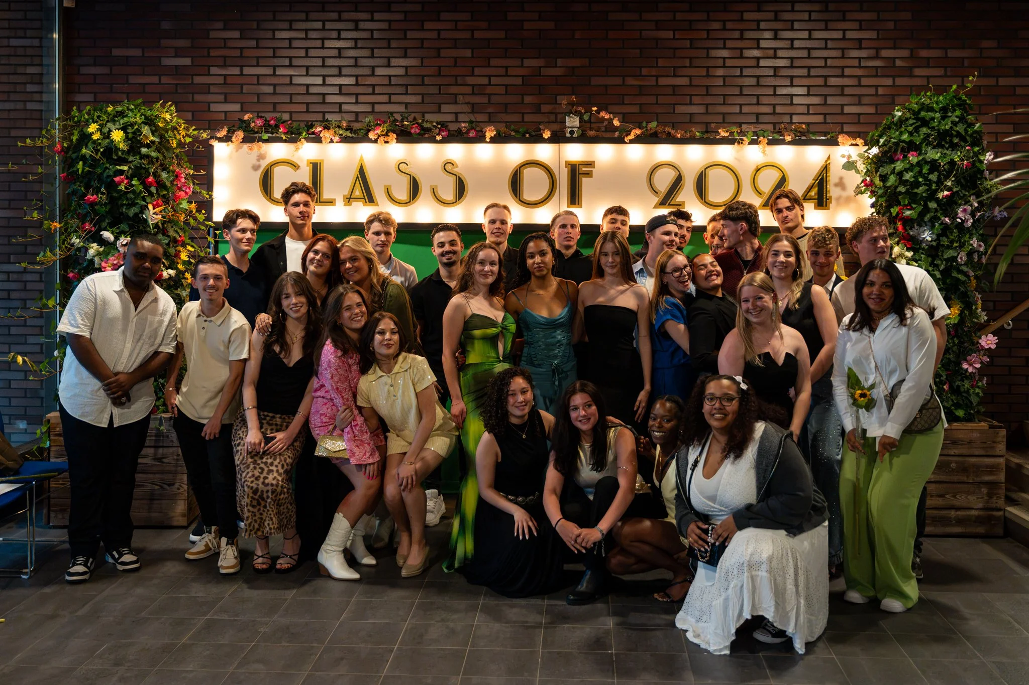 Een grote groep mensen die poseren voor een foto tijdens een afstudeerceremonie met een bord dat 'Class of 2024' zegt, omringd door bloemen en een bakstenen muur.