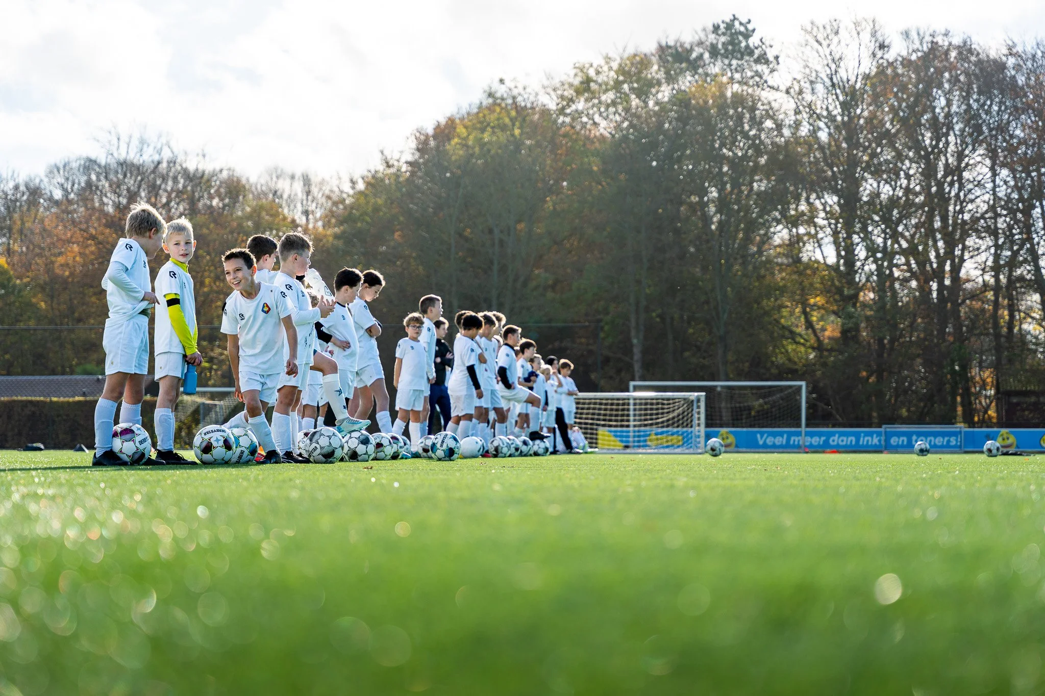 A group of young boys in white soccer uniforms standing on a green field, with soccer balls at their feet, preparing for a practice or game during daytime, with trees in the background.