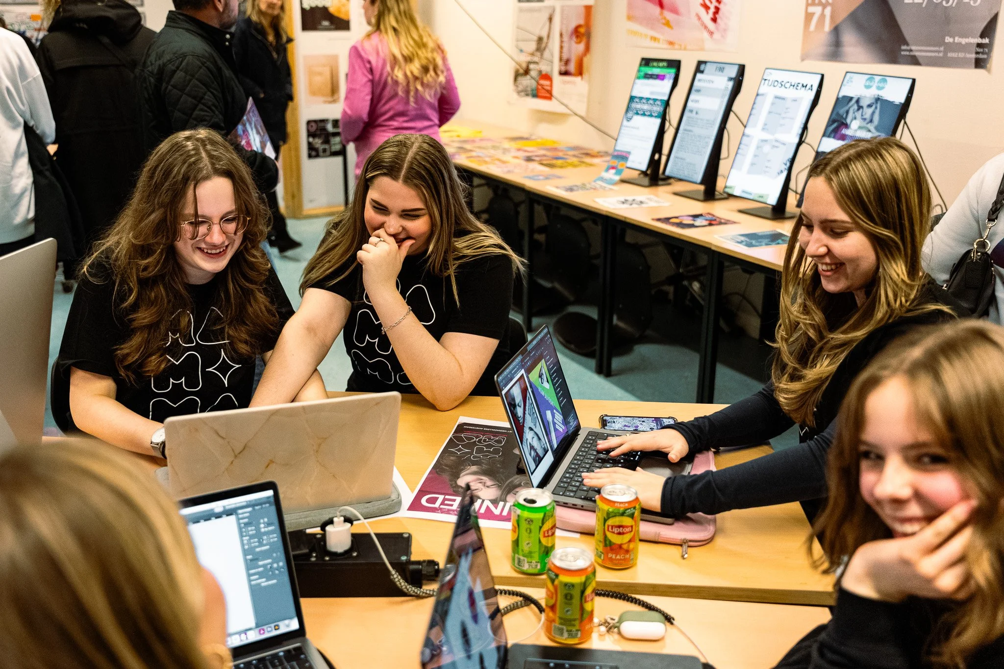 Group of young women sitting at a table with laptops, smiling and laughing during a tech event or workshop.