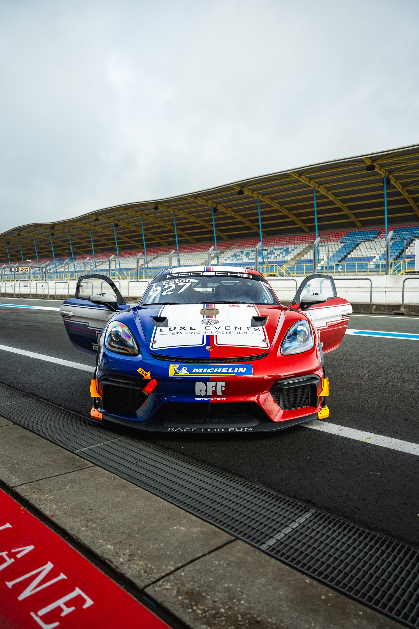 A colorful race car parked in the pit lane at a racetrack, with its doors open and a grandstand in the background under a cloudy sky.