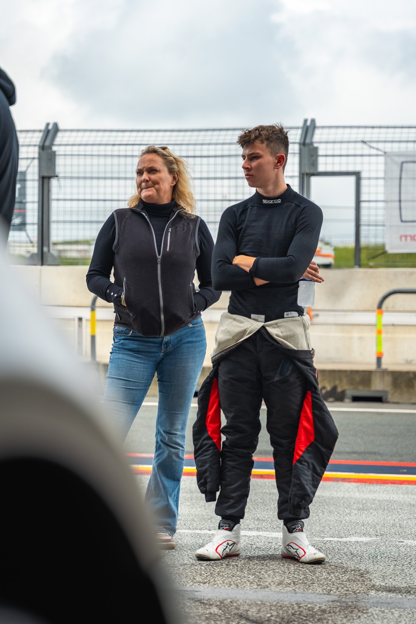 Twee mensen, een vrouw en een jongeman, staan samen op een racebaan onder een bewolkte hemel, gekleed in racekleding en leren schoenen.