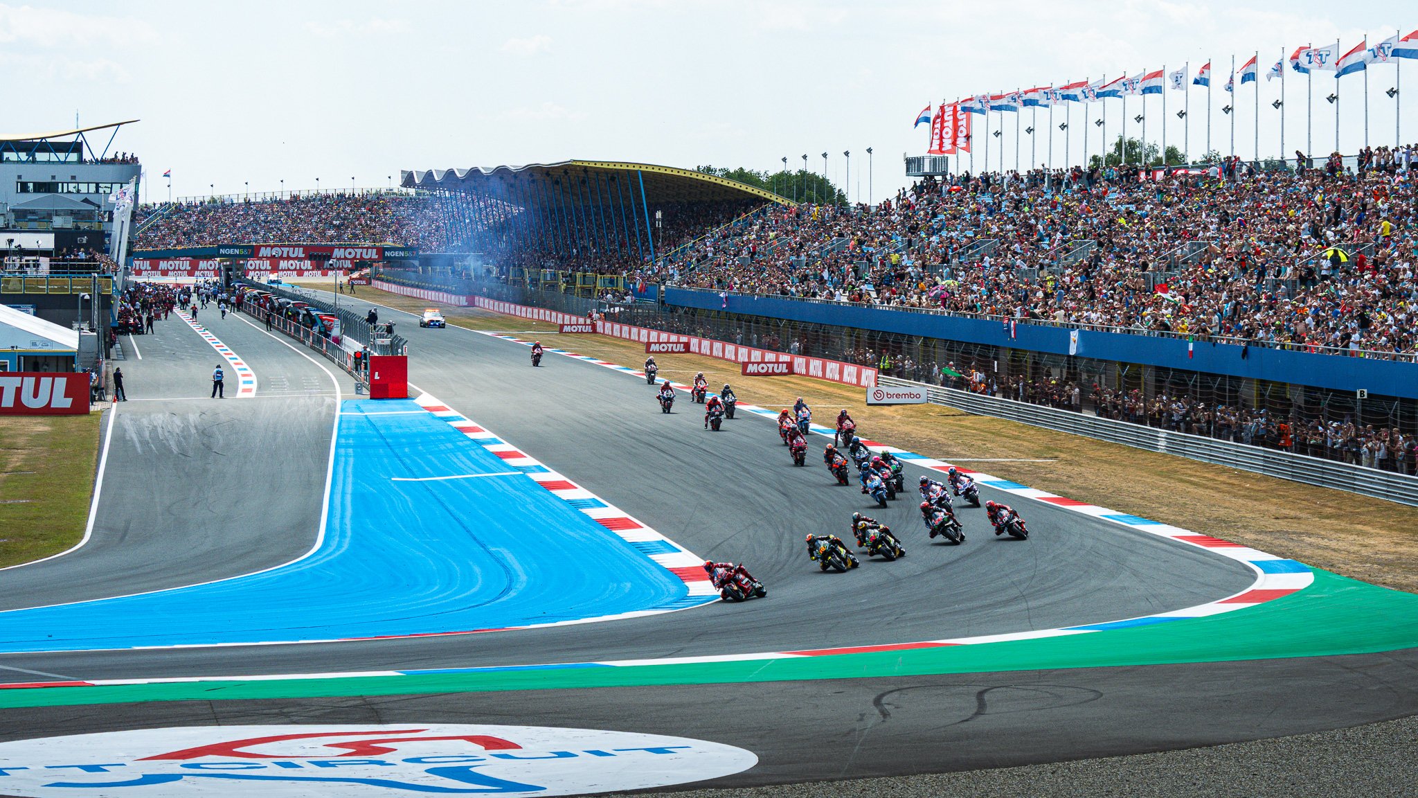 Motorcycle racers speeding around a racetrack with a large audience in the grandstands and flags flying in the background.