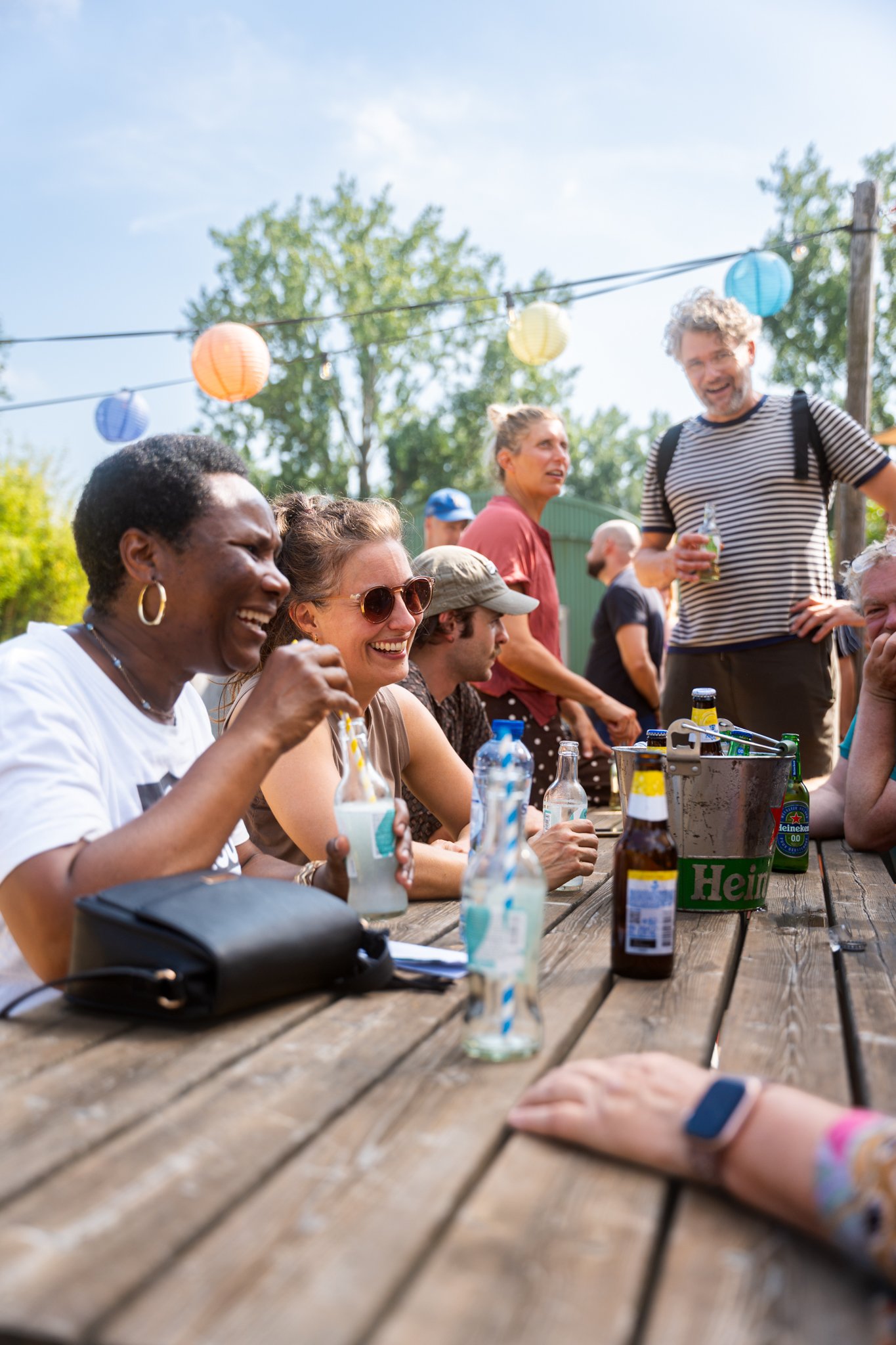 Een groep mensen zit aan een houten buitentafel, lachen en drinken, met kleurrijke lampions in de achtergrond op een zonnige dag.