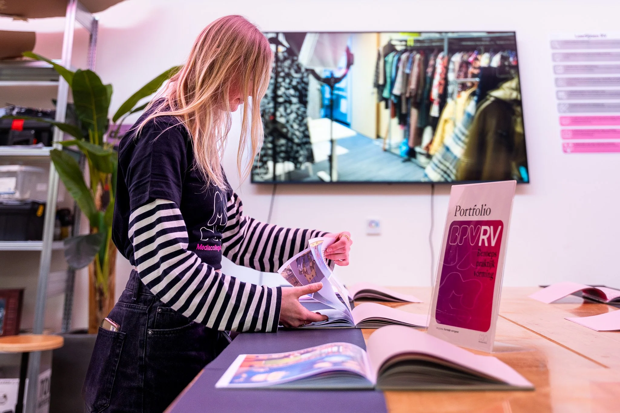 A young blonde woman browsing through photo albums inside a store, with shelves and a TV displaying clothing in the background.