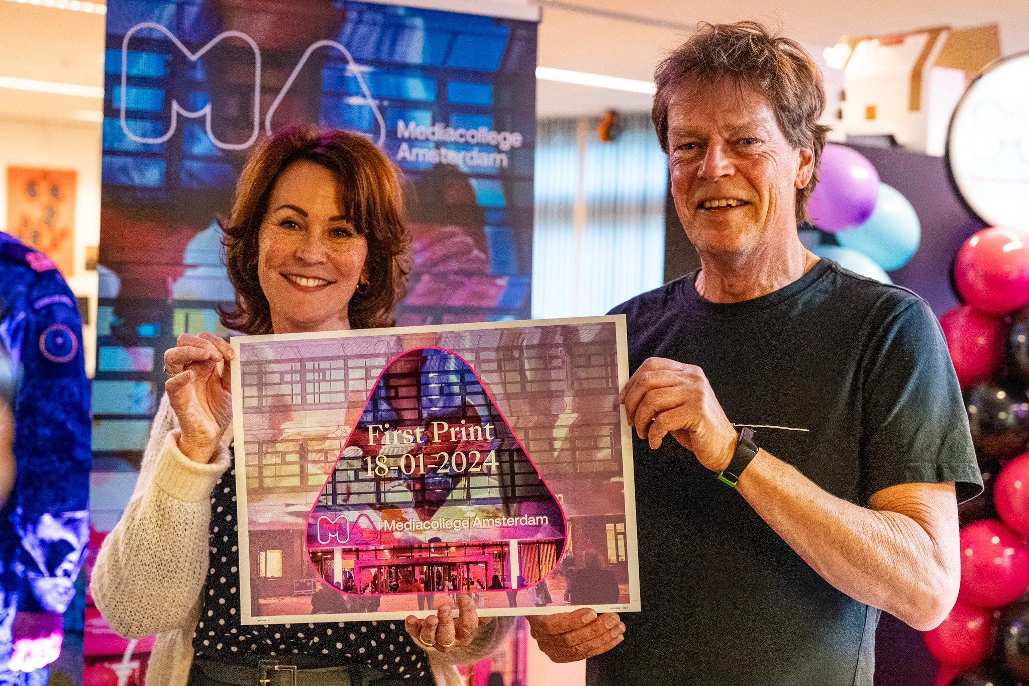 Two people smiling and holding a sign that reads "First Print 18-01-2024 Mediagallege Amsterdam" in front of a colorful background with balloons and a poster.