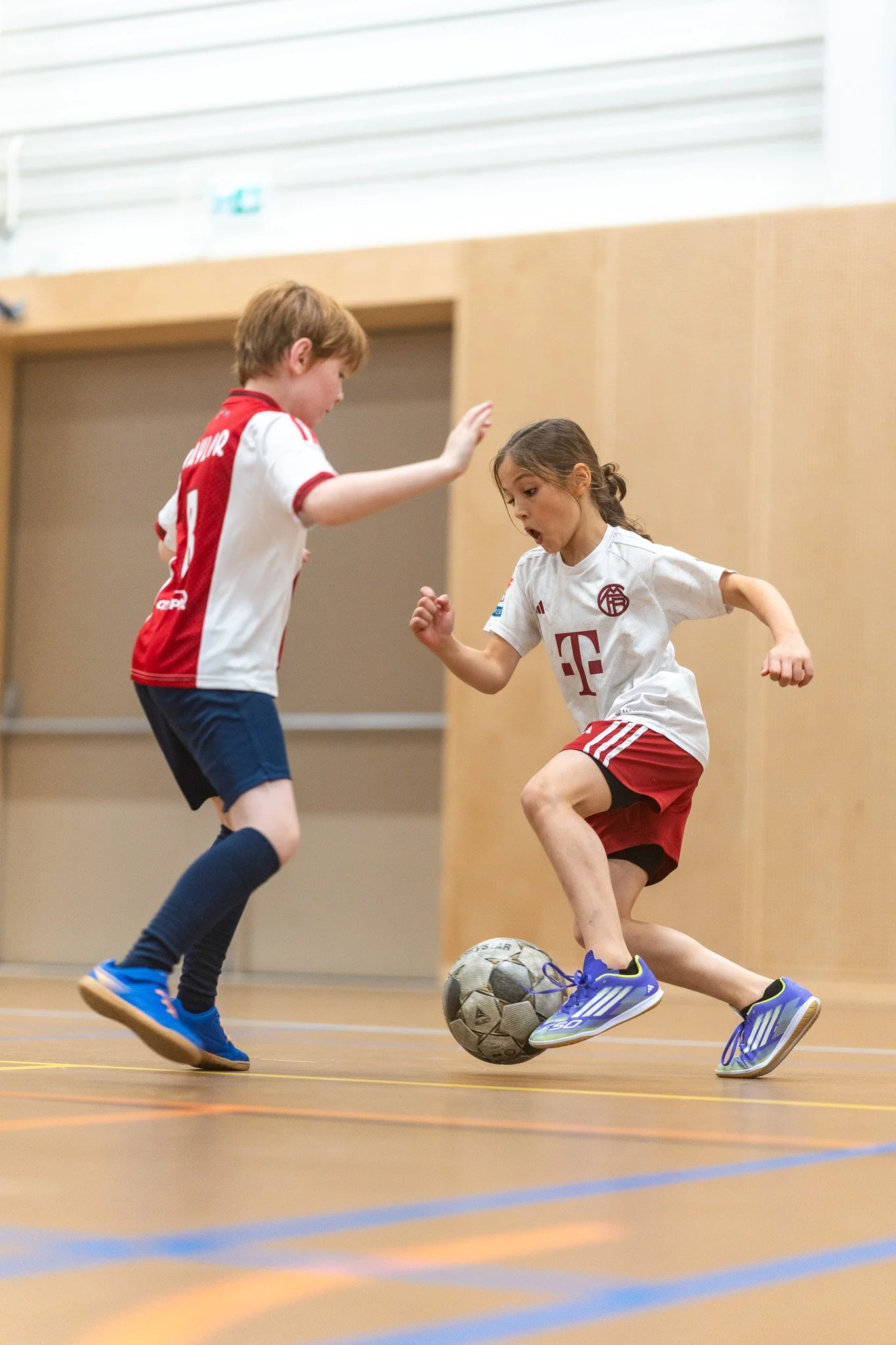 Two young children playing indoor soccer; one girl in a white and red jersey controlling a ball while another boy in a red and white jersey approaches to challenge.