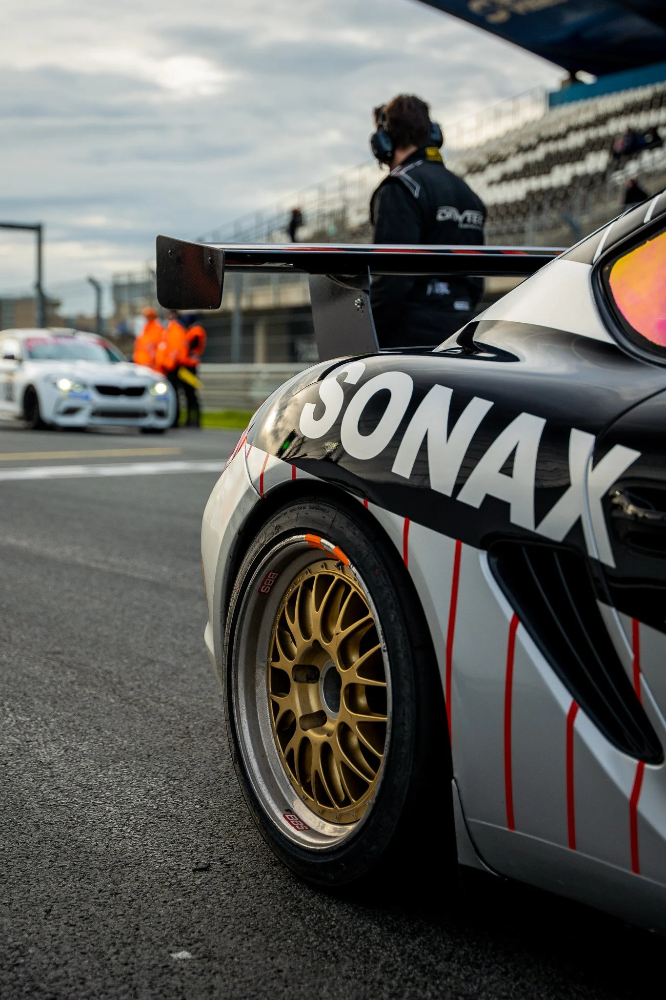 Close-up of a racing car with a black and white body and gold wheels, showing the name 'SONAX' on the side. In the background, a person with headphones and an event staff in orange are near a white race car on a race track under a cloudy sky.