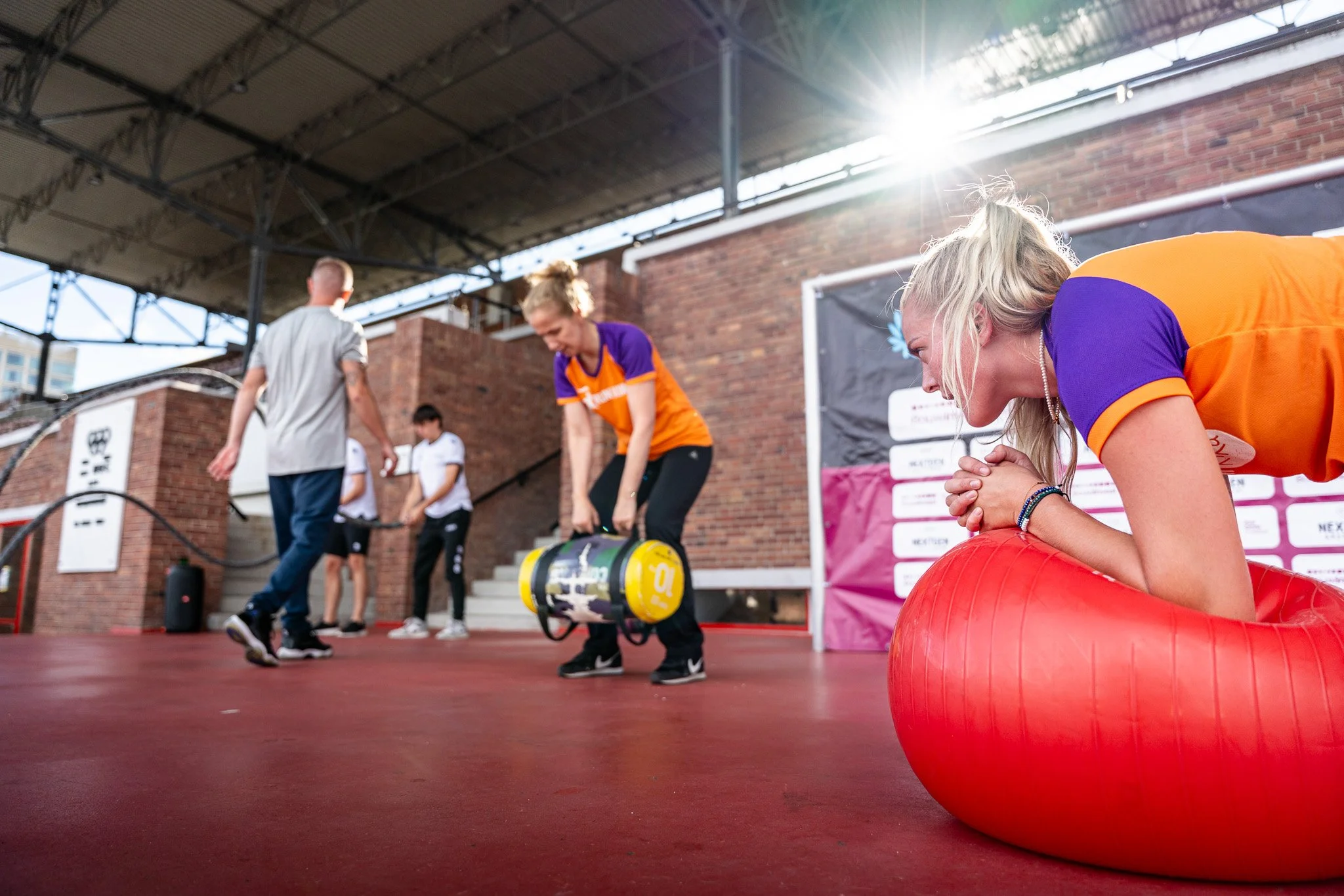 People exercising on a stage, with some stretching and others preparing for a workout, under a canopy with sunlight.