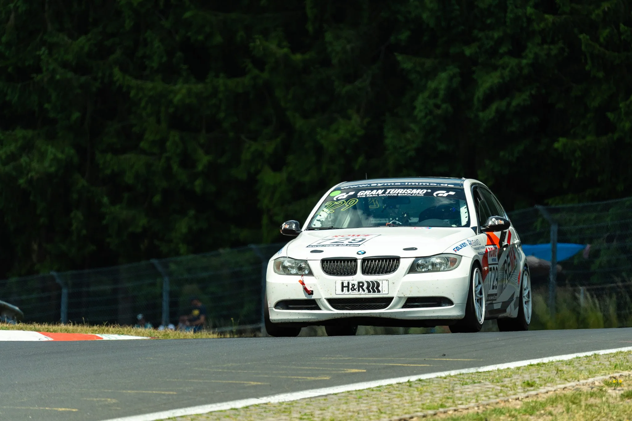 Witte raceauto met sponsorstickers rijdt snel op een circuit, omringd door bomen.