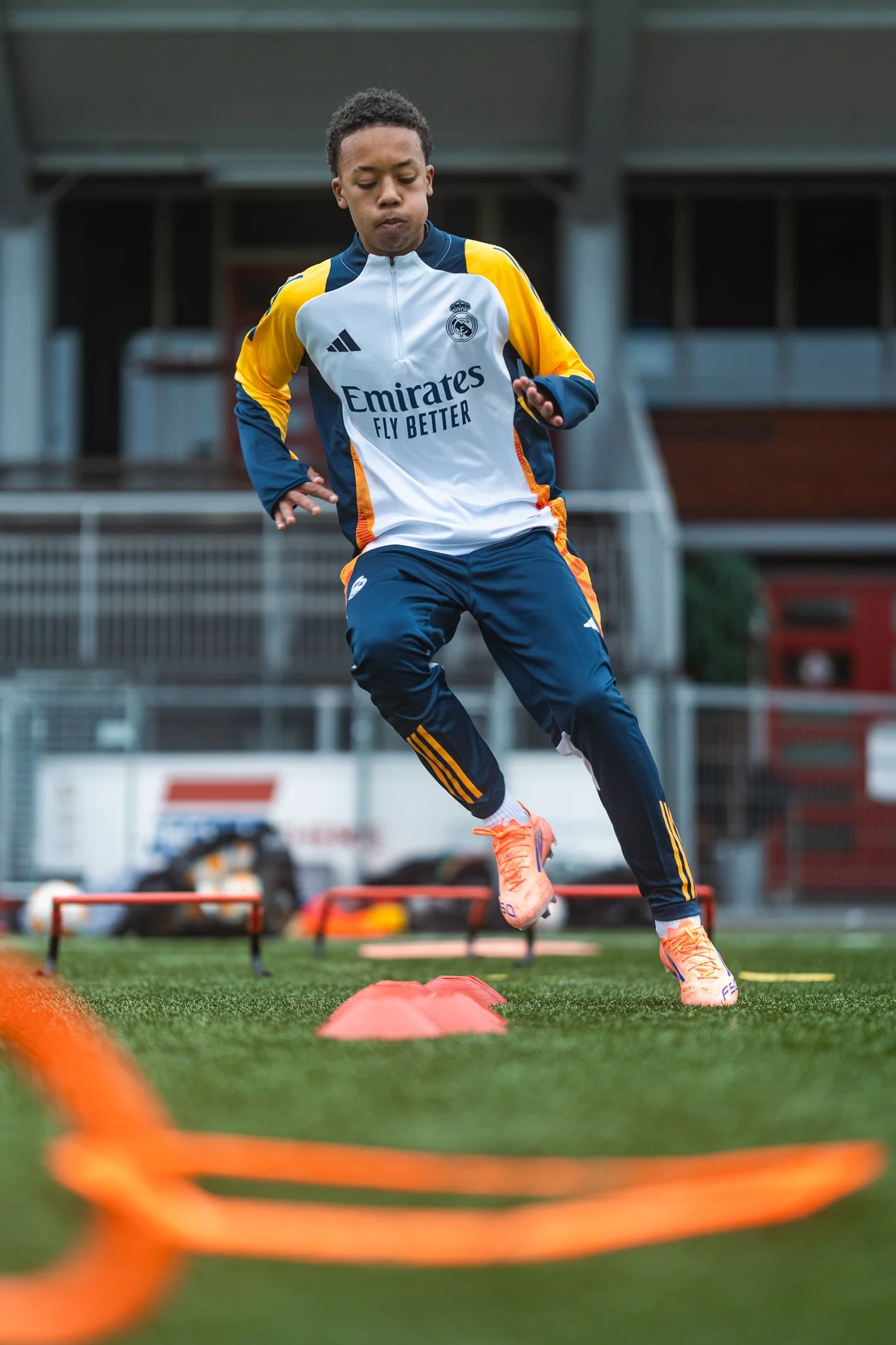 een jongen op een sportveld tijdens trainingen, hij draagt een sportoutfit met het Real Madrid logo, hij springt over een hindernis met andere trainingsmaterialen op de grond.
