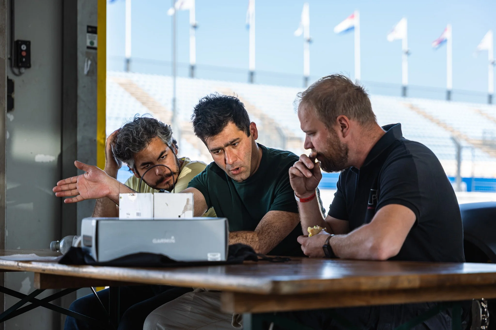 Drie mannen zitten samen aan een tafel, verdiept in een gesprek of onderzoek, met een achtergrond van vlaggen en een structuur van een stadion of tribune.