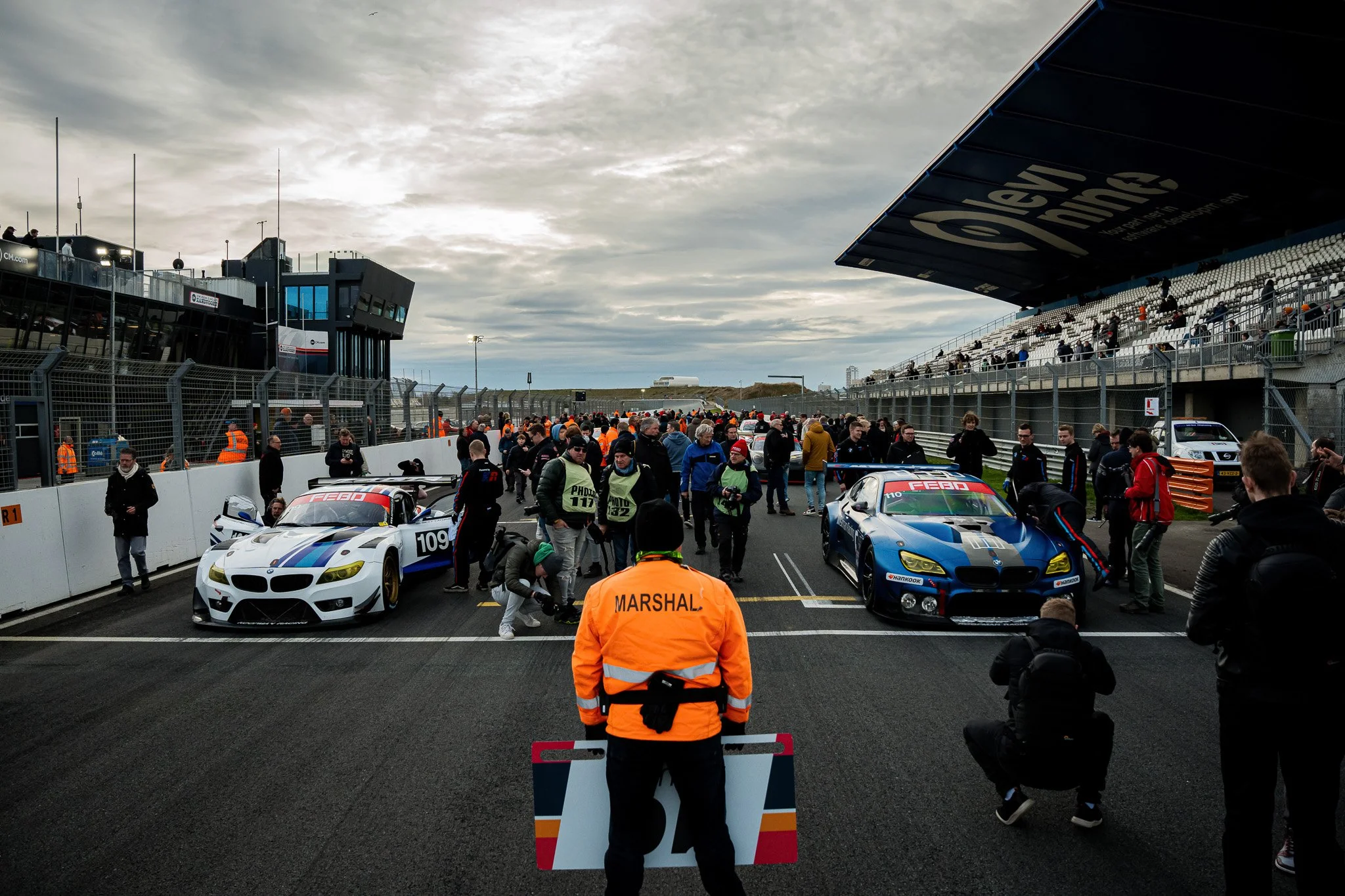 Een racebaan met sportwagens klaar voor een race, omringd door mensen en een haven met tribunes op de achtergrond. Er is een persoon met een oranje jas met de tekst 'Marshal' en een bord met het nummer 7.