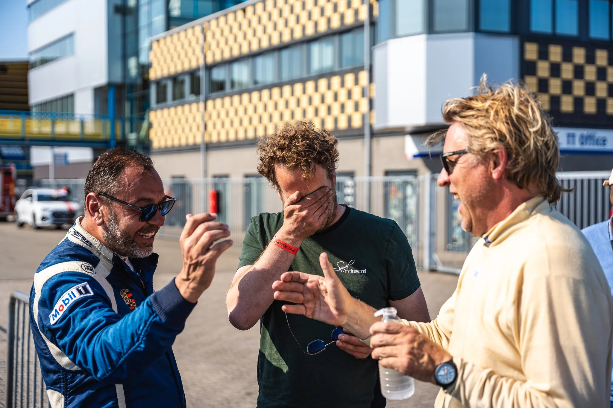 Drie mannen lachen en praten buiten tegen een achtergrond van een modern gebouw op een zonnige dag.