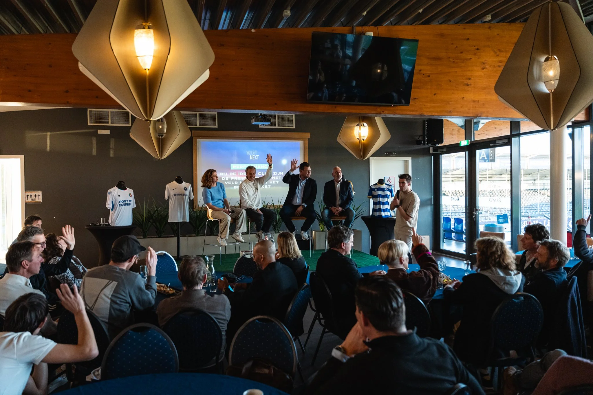 Binnenruimte met mensen die deelnemen aan een bijeenkomst, met panel op een verhoogd platform en display met tekst op de muur, versierd met voetbalshirts.