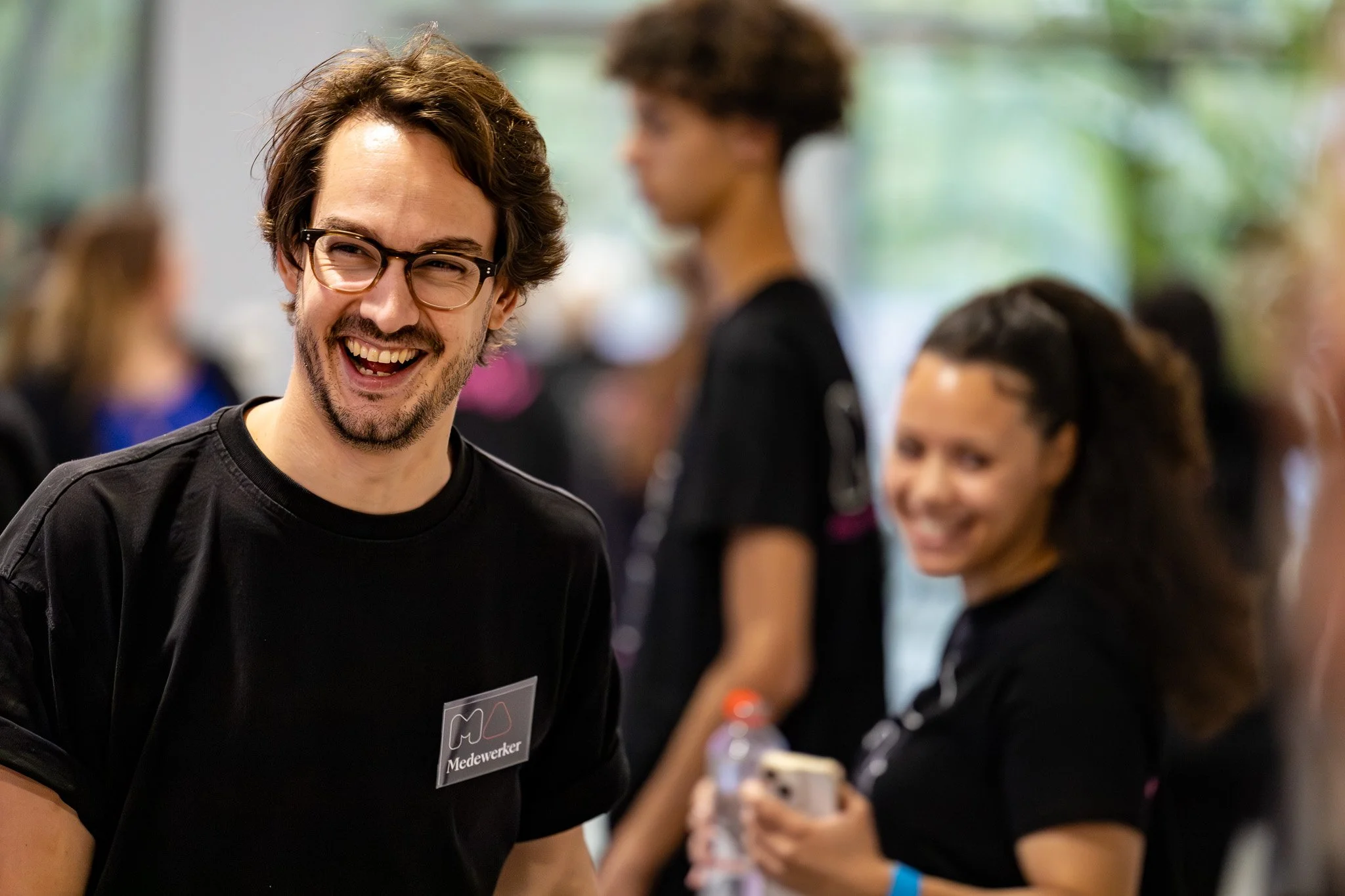 Young man with glasses, beard, and dark hair smiling, wearing a black shirt, at a social event. Woman in background with curly hair and a black shirt, holding a water bottle and smartphone, smiling.