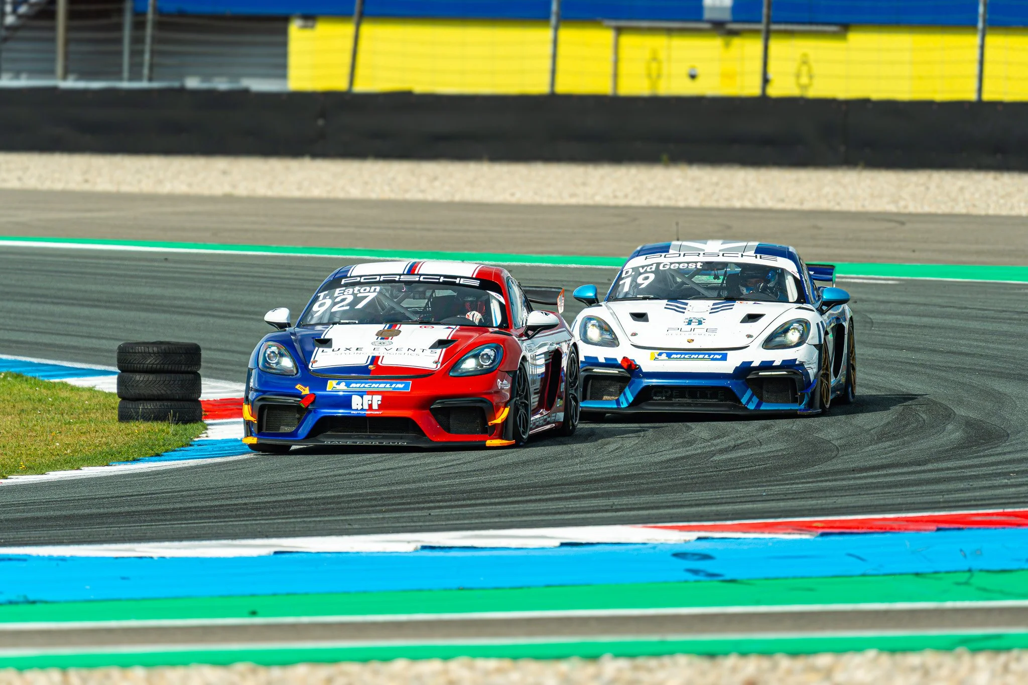 Twee raceauto's, een witte Porsche en een blauwe Porsche, rijden op een racebaan met gekleurde markeringen, omringd door een tribune en banden.
