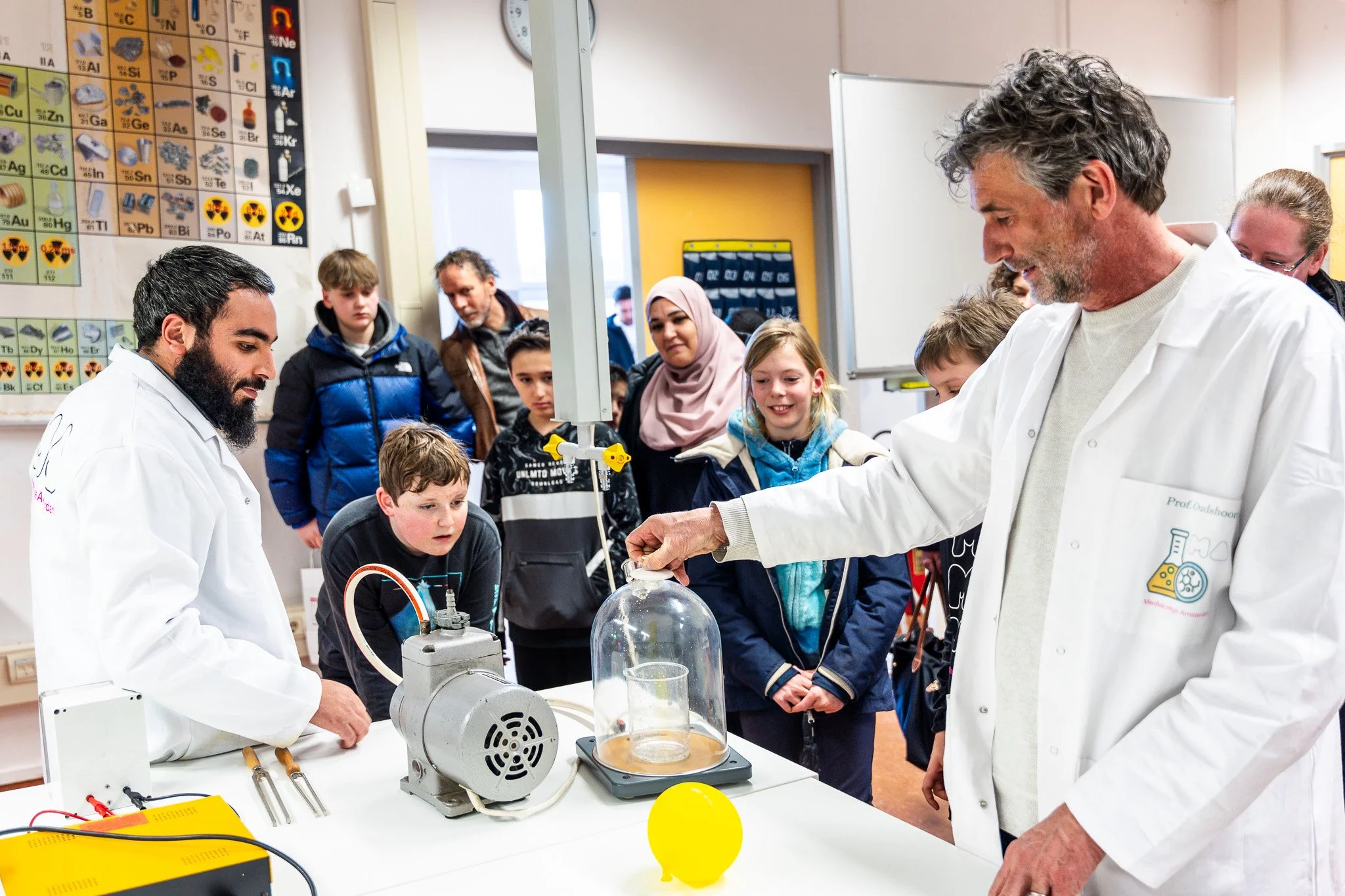 Students and teachers conduct a science experiment in a classroom with a periodic table poster on the wall.