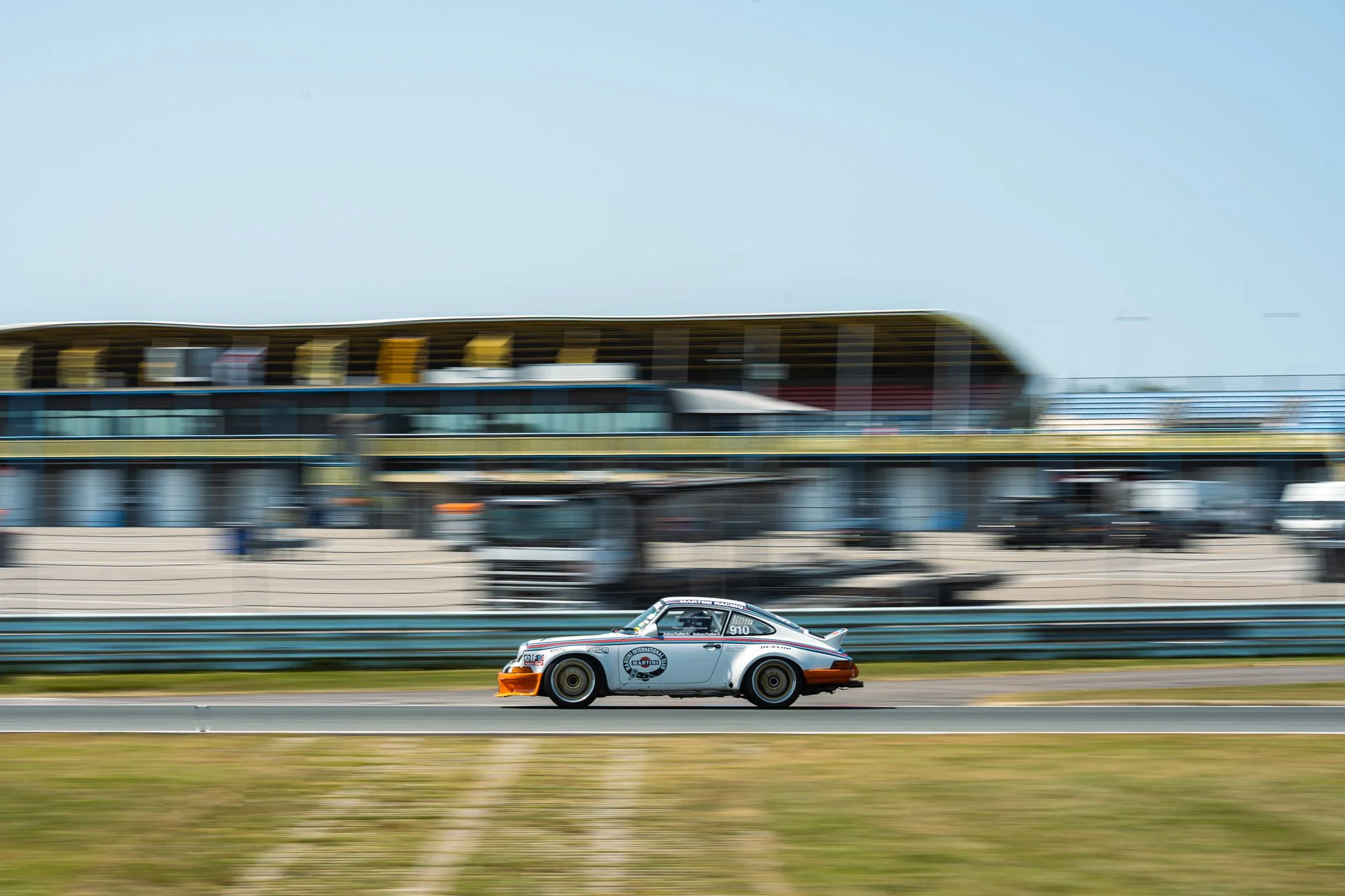 Een raceauto rijdt op een circuit, met een modern gebouw op de achtergrond. De auto is wit met oranje accenten en logo's, en beweegt snel over de baan.