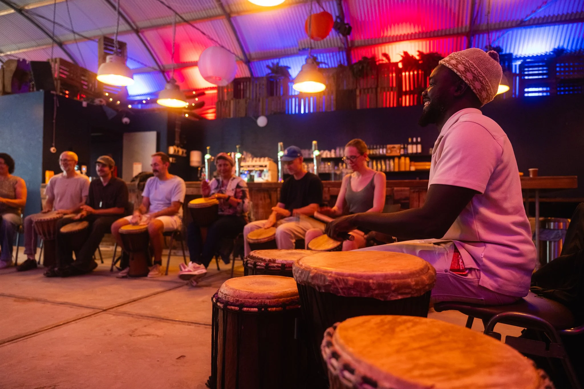 Man speelt op djembé drum tijdens een bijeenkomst, met mensen die ernaast zitten, binnen een kleurrijk verlichte ruimte.