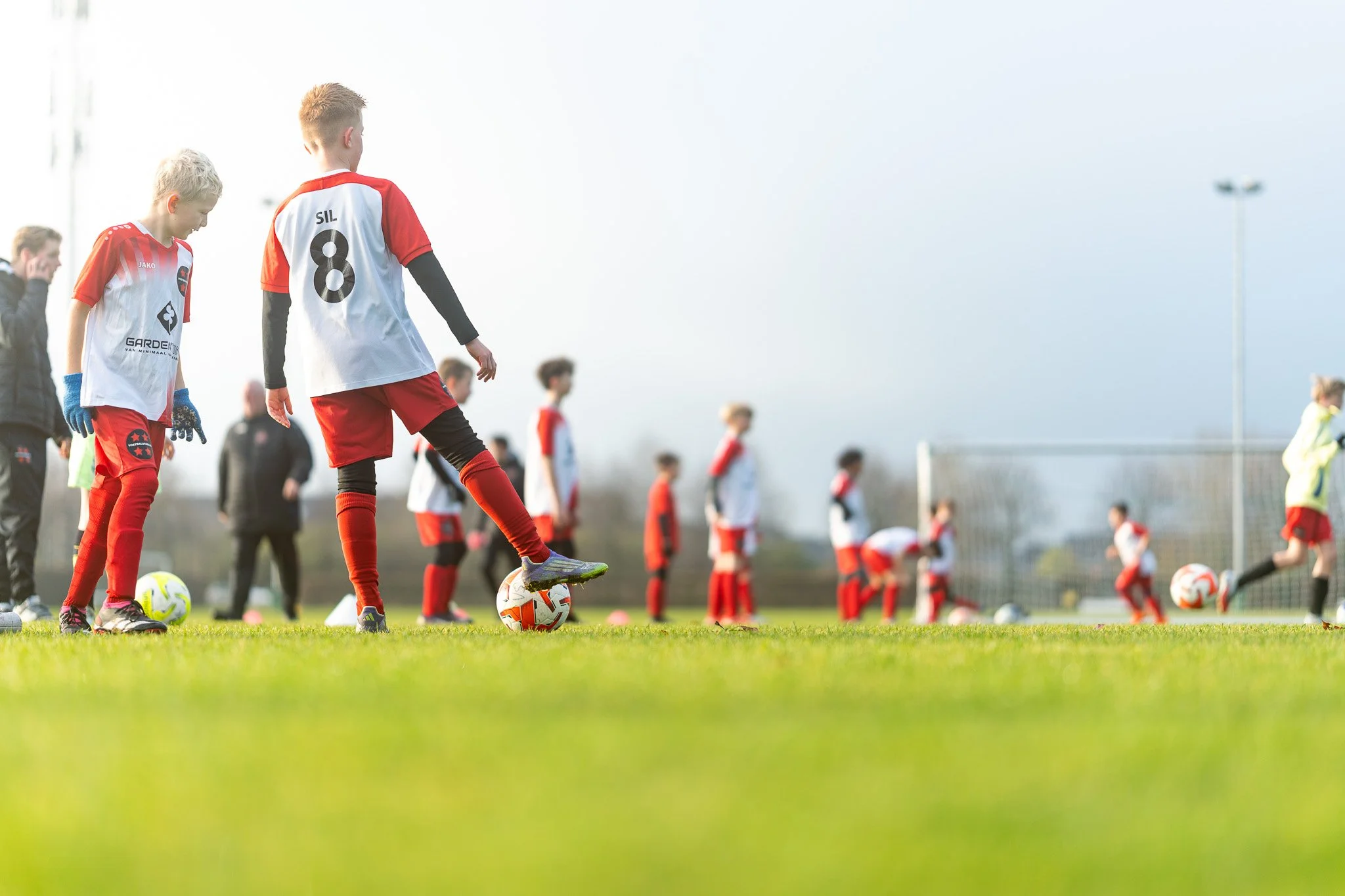Young boys wearing red and white soccer uniforms practice on a grassy field, with several soccer balls and a goalpost in the background.