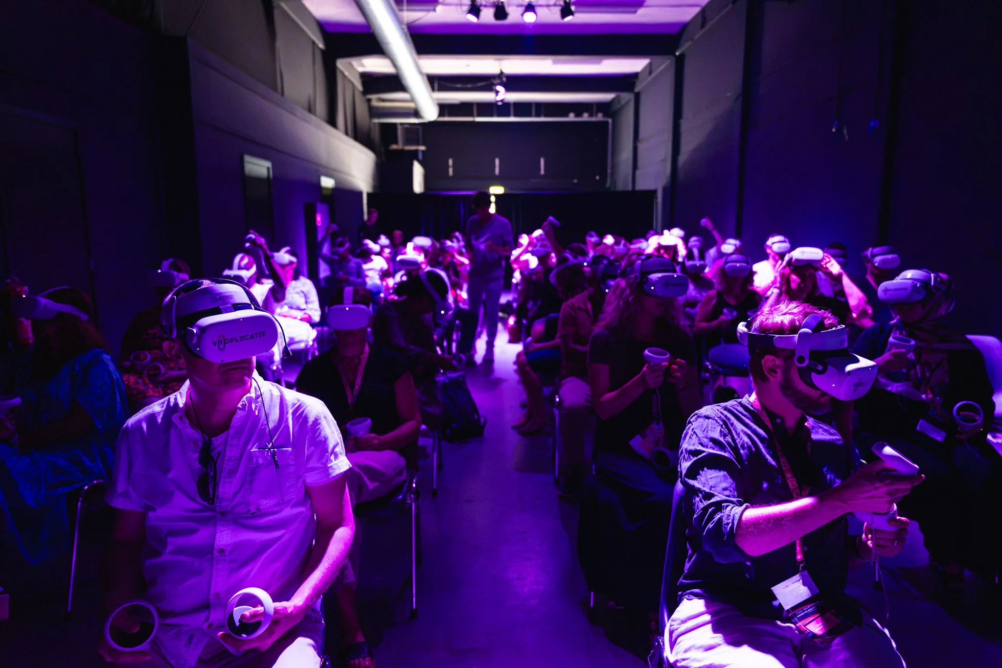 Groep mensen die virtual reality-headsets dragen tijdens een evenement of conferentie, zit in een donkere ruimte met paarse verlichting.