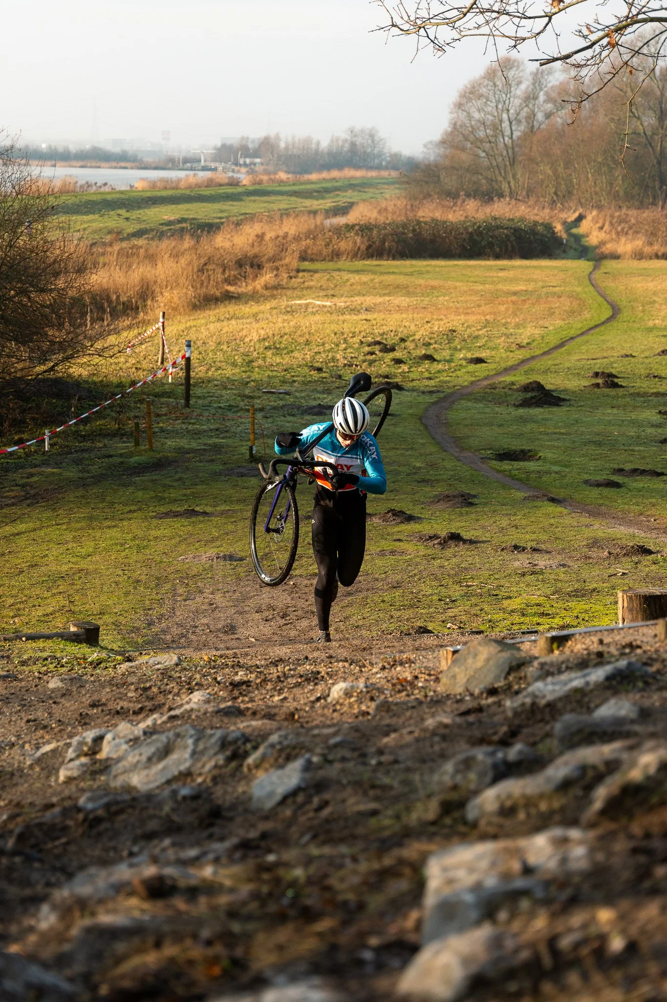 A cyclist carrying their bike while running up a rugged outdoor trail in a grassy park with a river in the background.