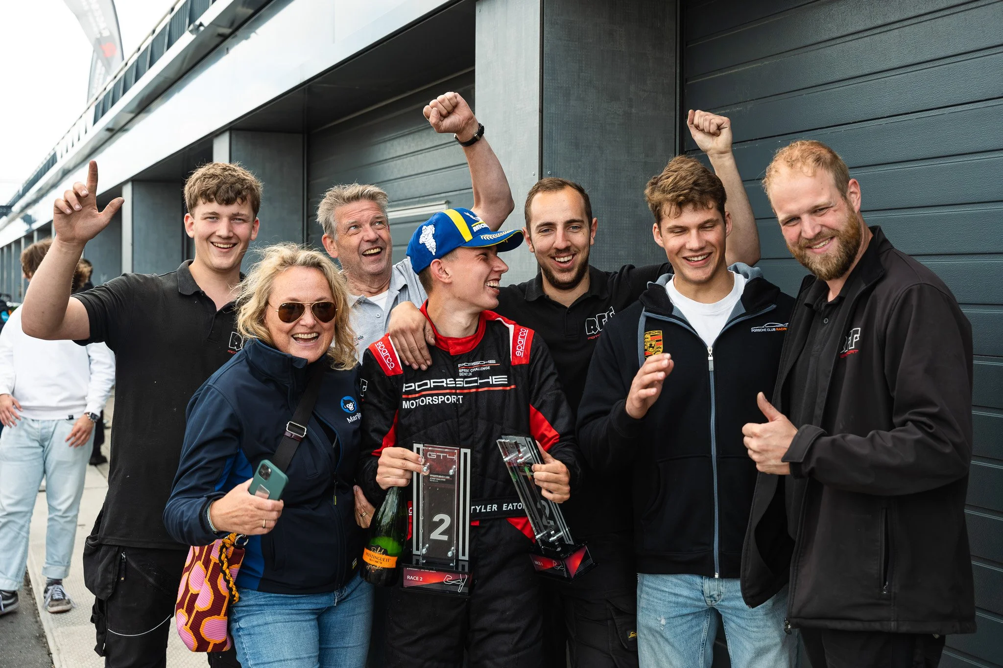 Groep mensen, inclusief een coureur in racepak, vieren een overwinning met champagne en bekers buiten een gebouw. Ze lachen en juichen, gezonde sfeer van triomf.