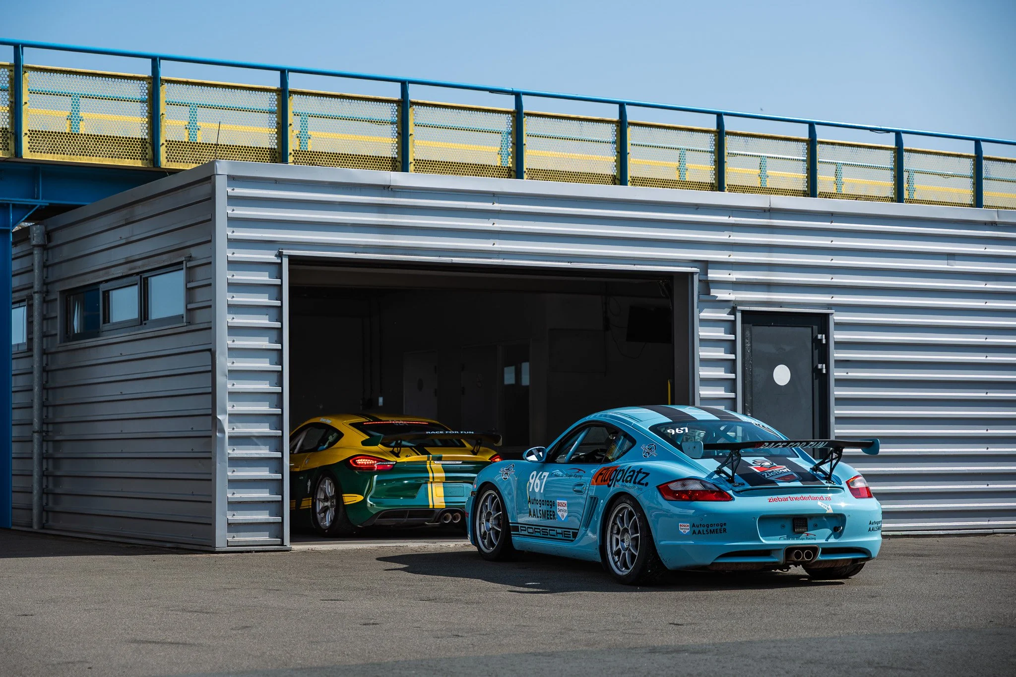 Two race cars, a yellow and green one and a light blue one, parked inside a garage at a racing track. The blue car has a large rear wing and race stickers.