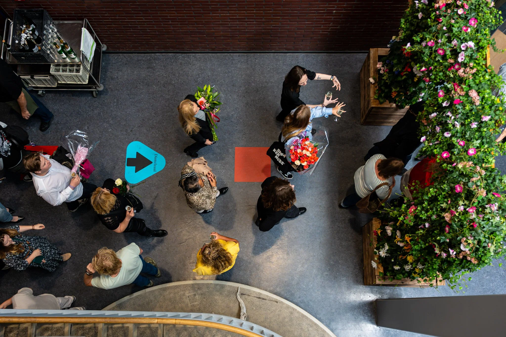 Bovenaanzicht van een groep mensen in een hal, met bloemen en een wit en groen bord die naar rechts wijzen.