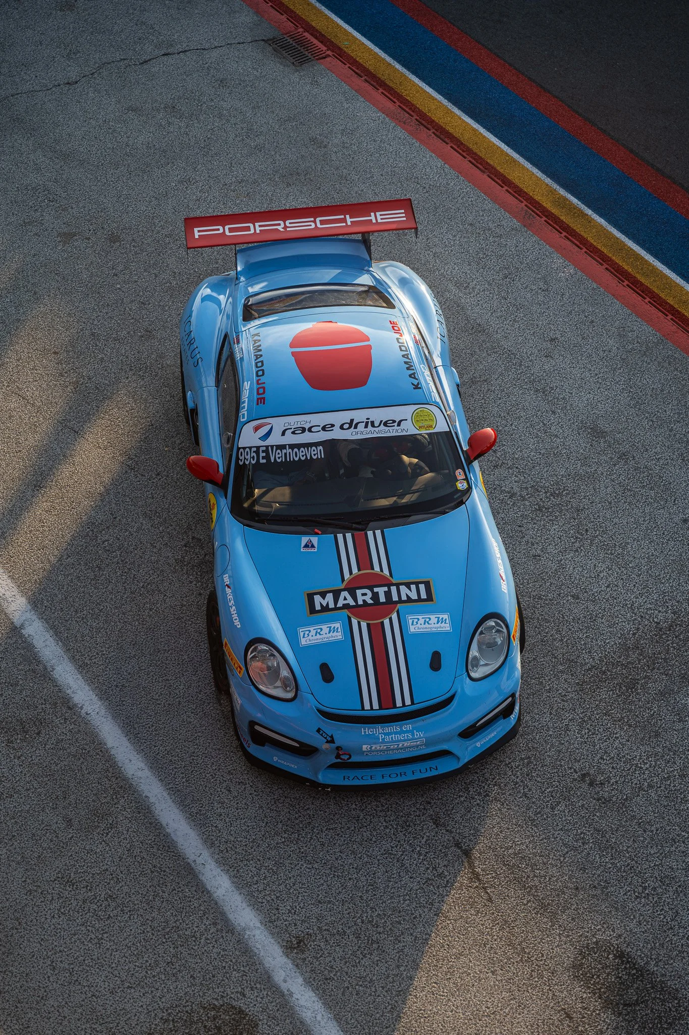 Een blauwe raceauto met Porsche- en Martini-logo's, geparkeerd op een asfaltweg met kleurrijke strepen aan de rand.