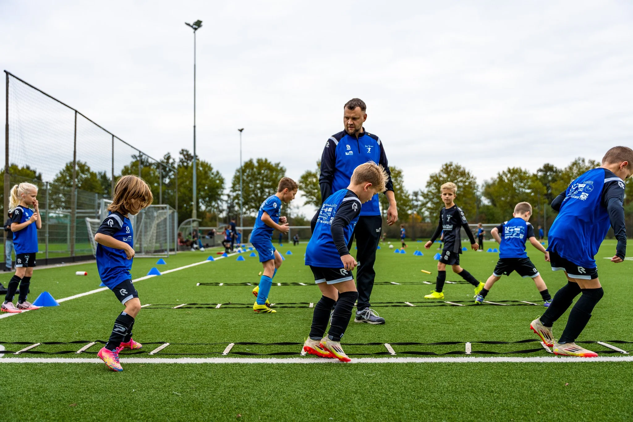 Een groep kinderen en een coach die op een grasveld trainen met agility-verloopmaterialen, zoals ladders en kegels, onder een bewolkte hemel.