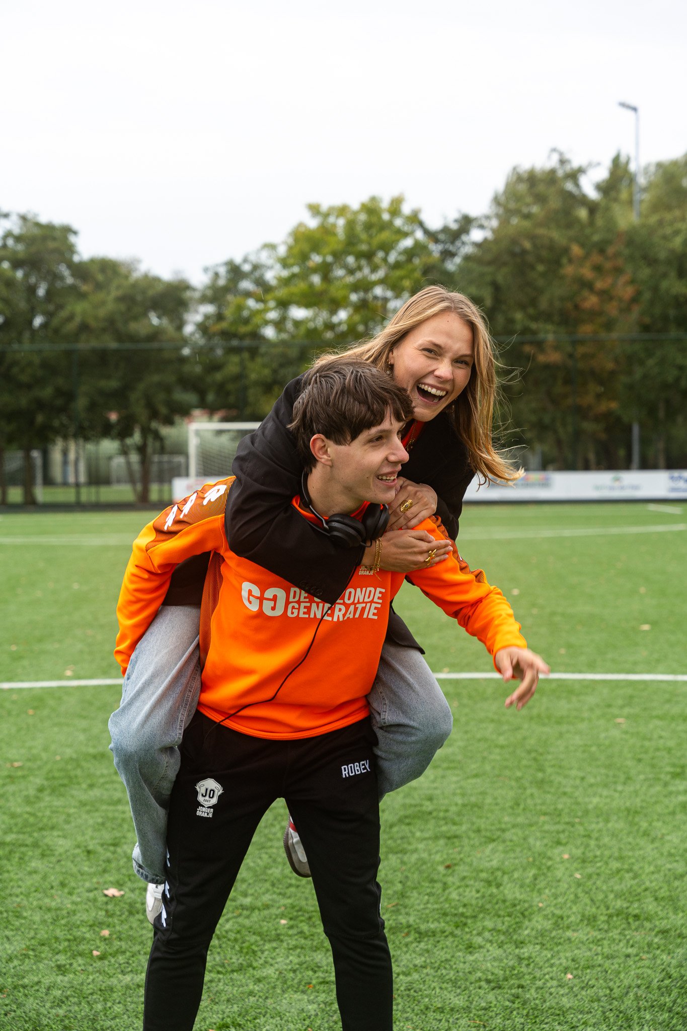 A woman giving a piggyback ride to a young man on a soccer field, both smiling and laughing. The woman has long blonde hair, and the young man is wearing an orange sports jersey and black pants.